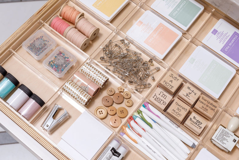 An assortment of neatly organized crafting supplies, including threads, beads, pins, buttons, stamps, and markers, in an acrylic drawer organizer.