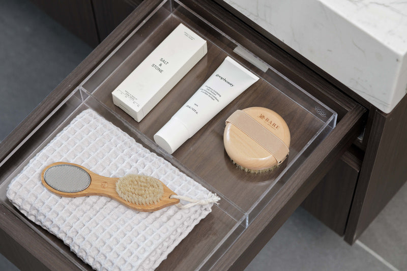 A clear acrylic bathroom organizer tray arranged in an open dark wood drawer, holding a white textured towel, a wooden-handled mirror, a white tube, a cream box, and a round beige makeup compact. A white marble countertop and dark cabinet are visible around it.