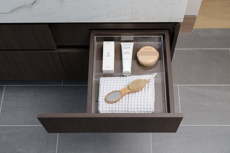 An open dark wood bathroom drawer on a gray tiled floor, containing a clear organizer tray with skincare items and a wooden hairbrush on a white towel.