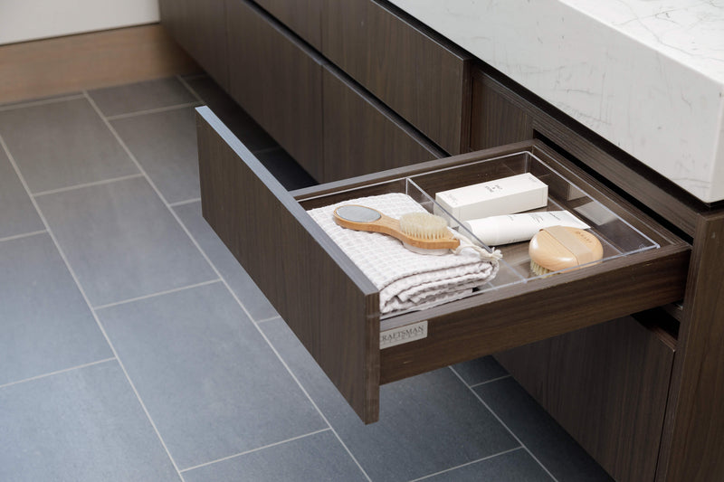 An open dark wood bathroom drawer with a clear organizer tray holding a folded towel, hairbrush, and small toiletries, installed under a white marble sink on a gray tiled floor.