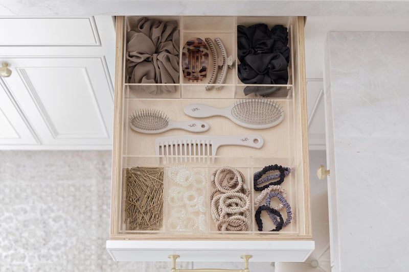 An open drawer with a custom acrylic Signature Drawer Organizer from Salt by Sabrina filled with hair accessories and hair tools, including scrunchies, hair ties, brushes, a comb, and clips, arranged neatly in a light wood compartment system.