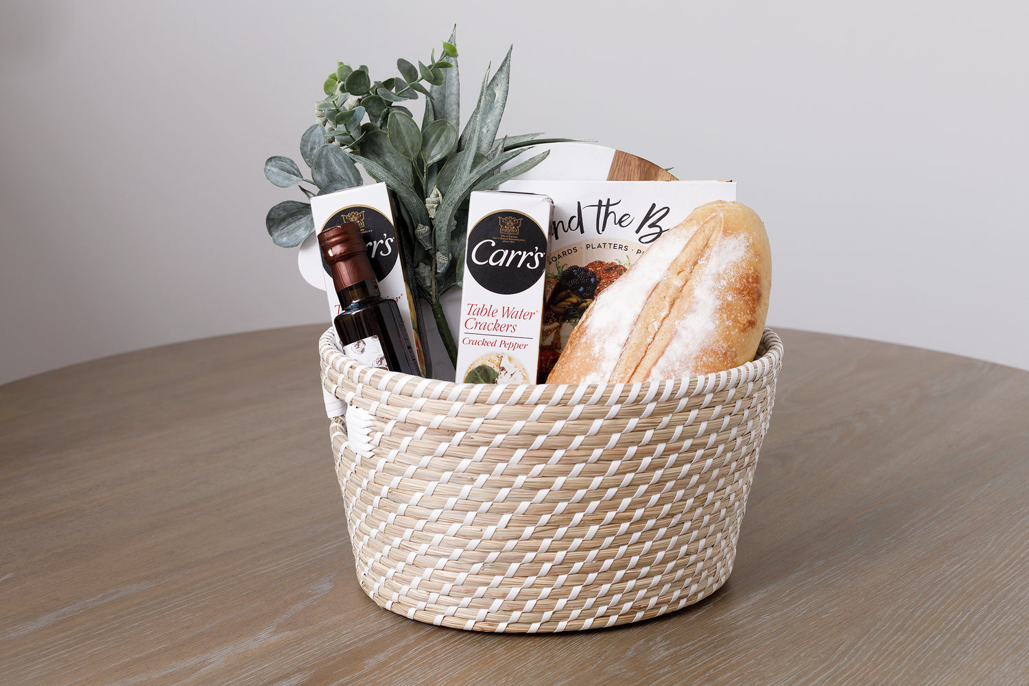Neutral-toned White Haven basket filled with goodies including fresh bread, olive oil and a cookbook.