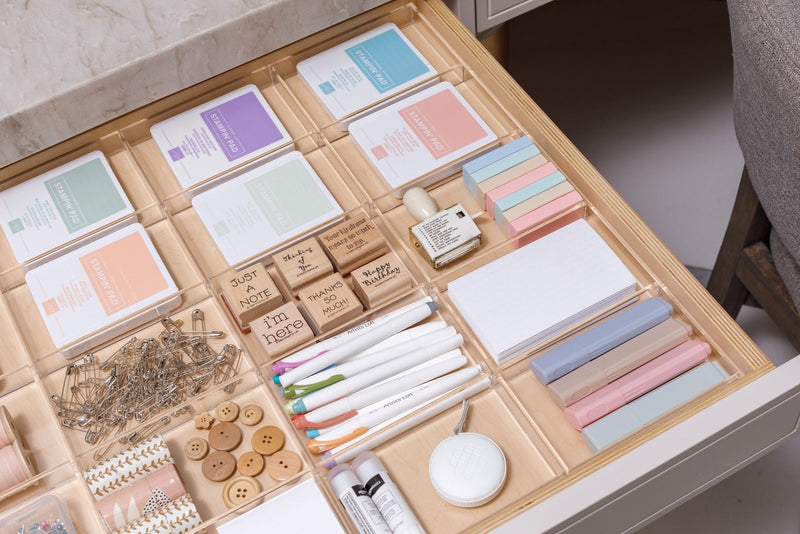 An assortment of neatly organized crafting supplies, including threads, beads, pins, buttons, stamps, and markers, in an acrylic drawer organizer.