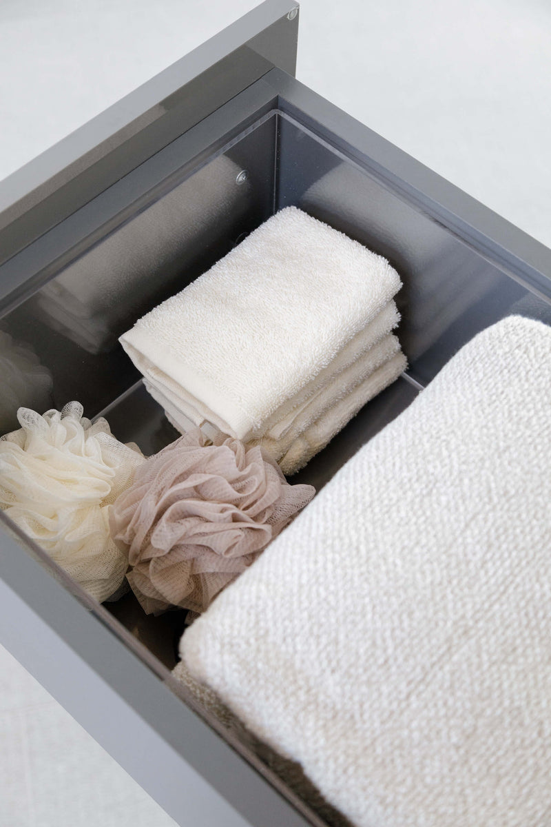 A close-up of a gray bathroom drawer containing folded white towels, a cream loofah, and a beige loofah.