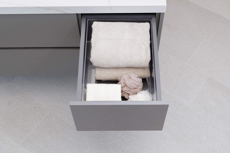 An open gray bathroom drawer containing folded towels, a bath sponge, and a loofah, set against a light gray tiled floor.