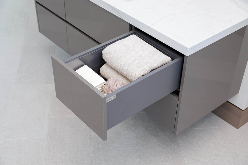 A modern bathroom vanity drawer with a gray exterior and white countertop, shown with one drawer pulled open to reveal folded towels and toiletries inside.